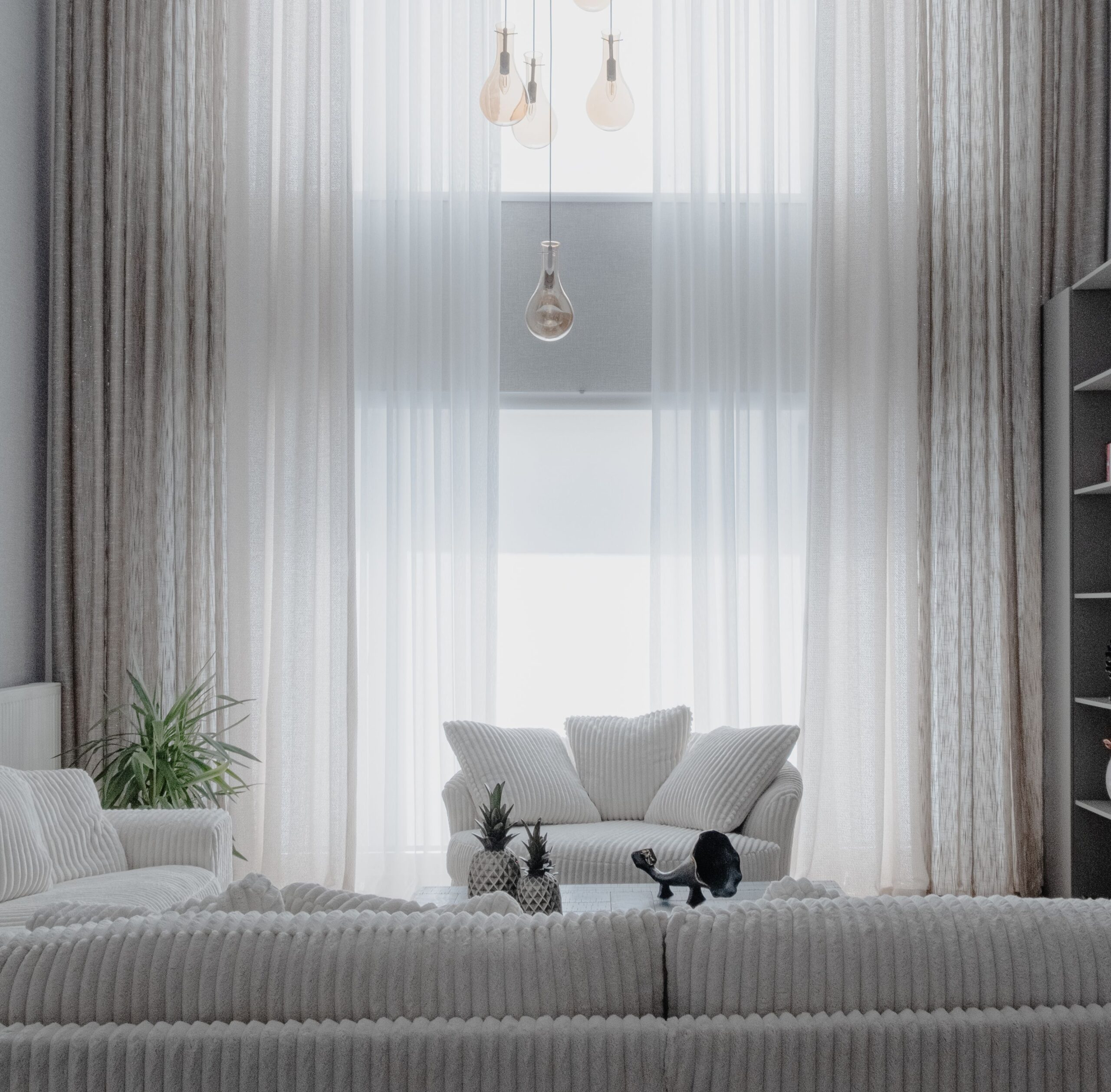 Cozy living room with white knitted sofa, cushions, a green plant, sheer curtains, and hanging light bulbs above.