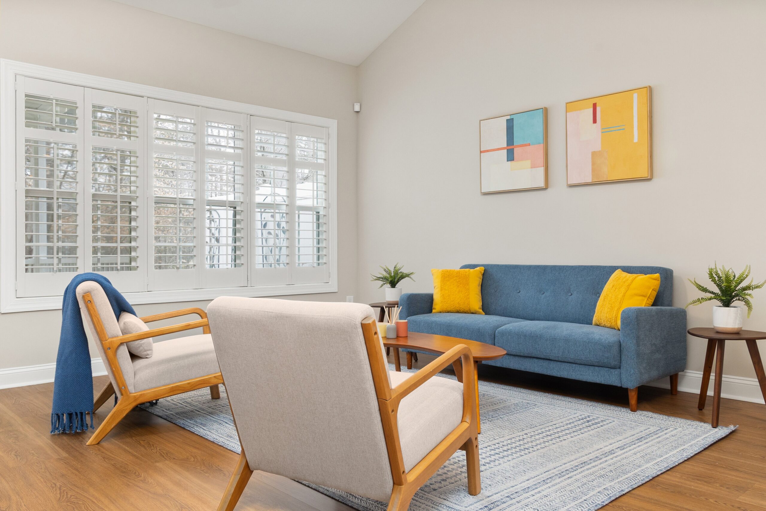 Bright living room with a blue sofa, yellow cushions, and light wood furniture beneath abstract wall art and large white shuttered windows.