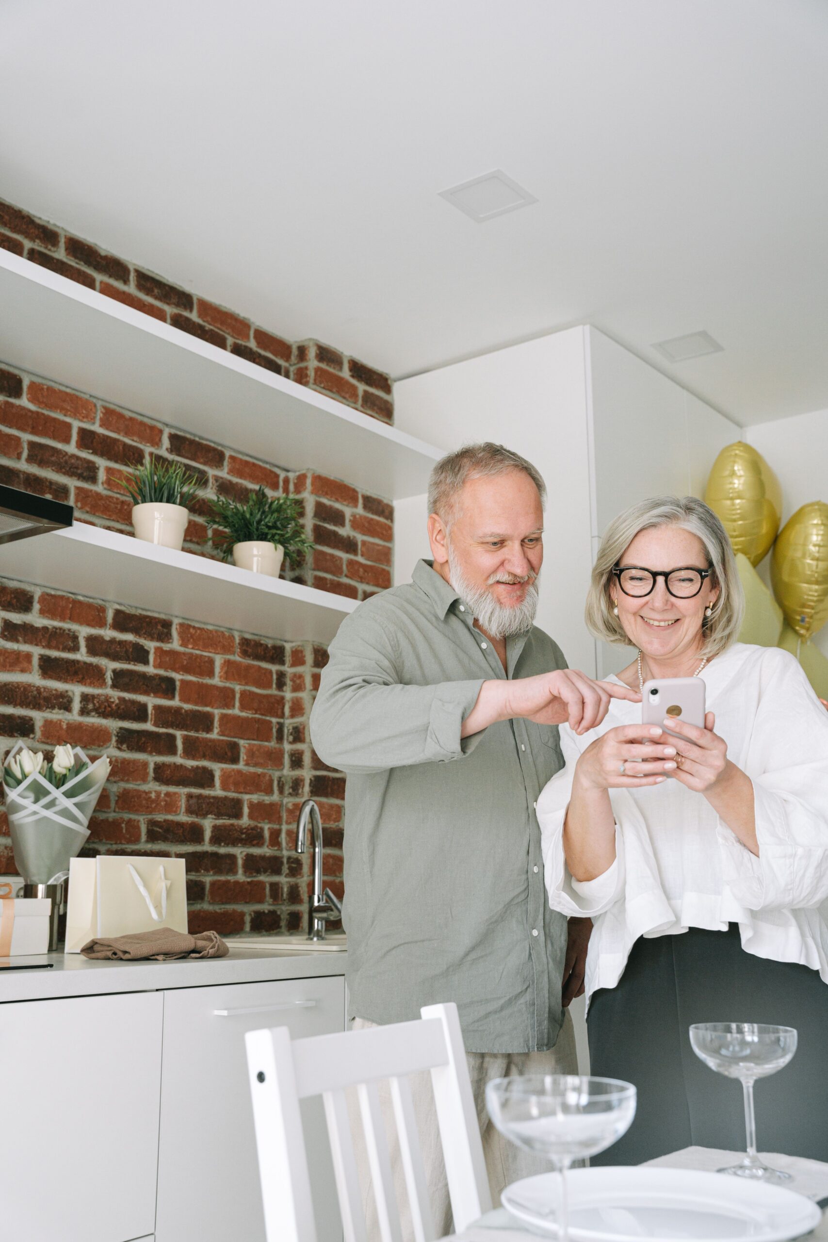 Couple in a bright kitchen looking at a smartphone together, with white cabinets and gold party balloons in the background