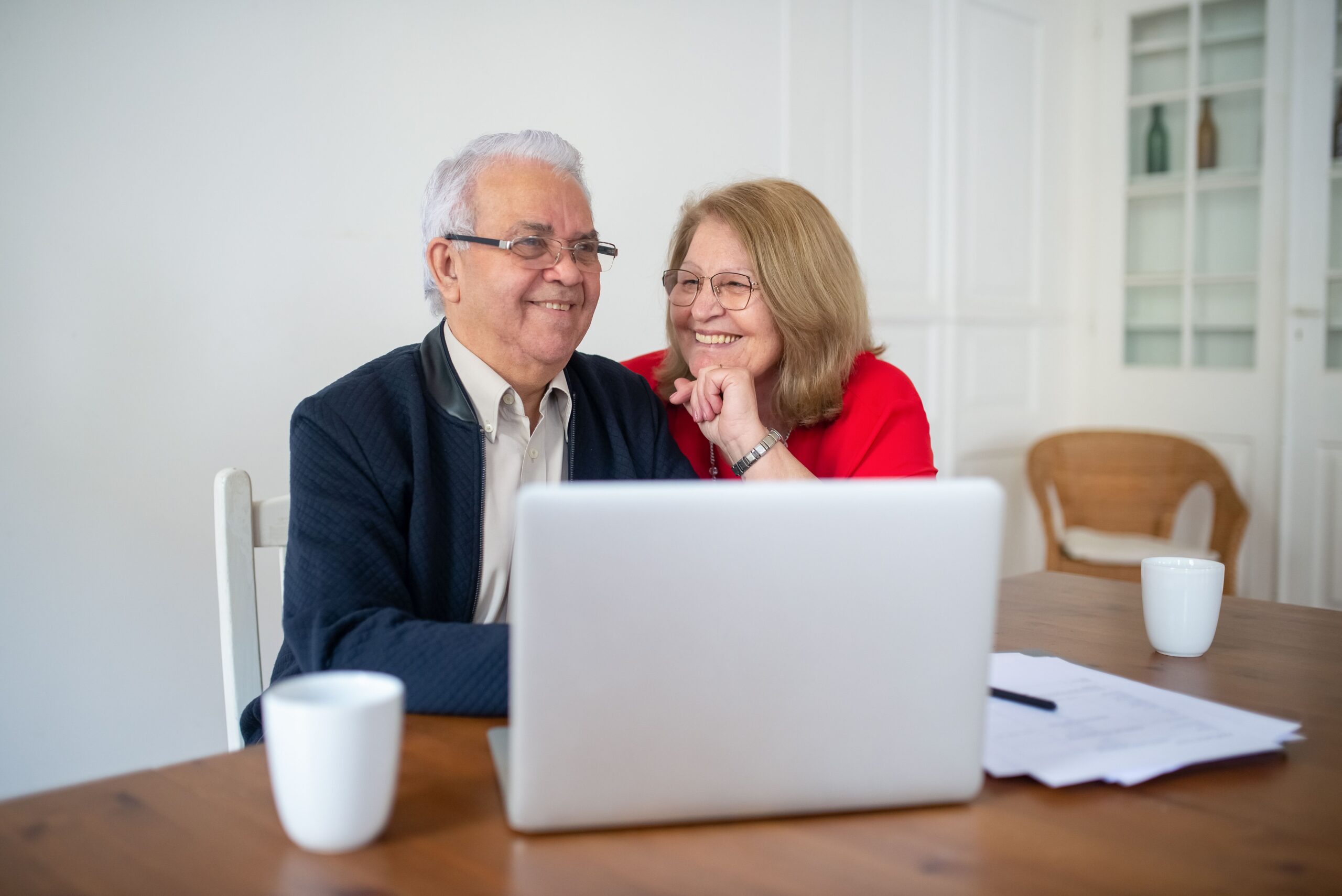 Two smiling older adults sit at a wooden table with a laptop, looking at each other in a bright room.
