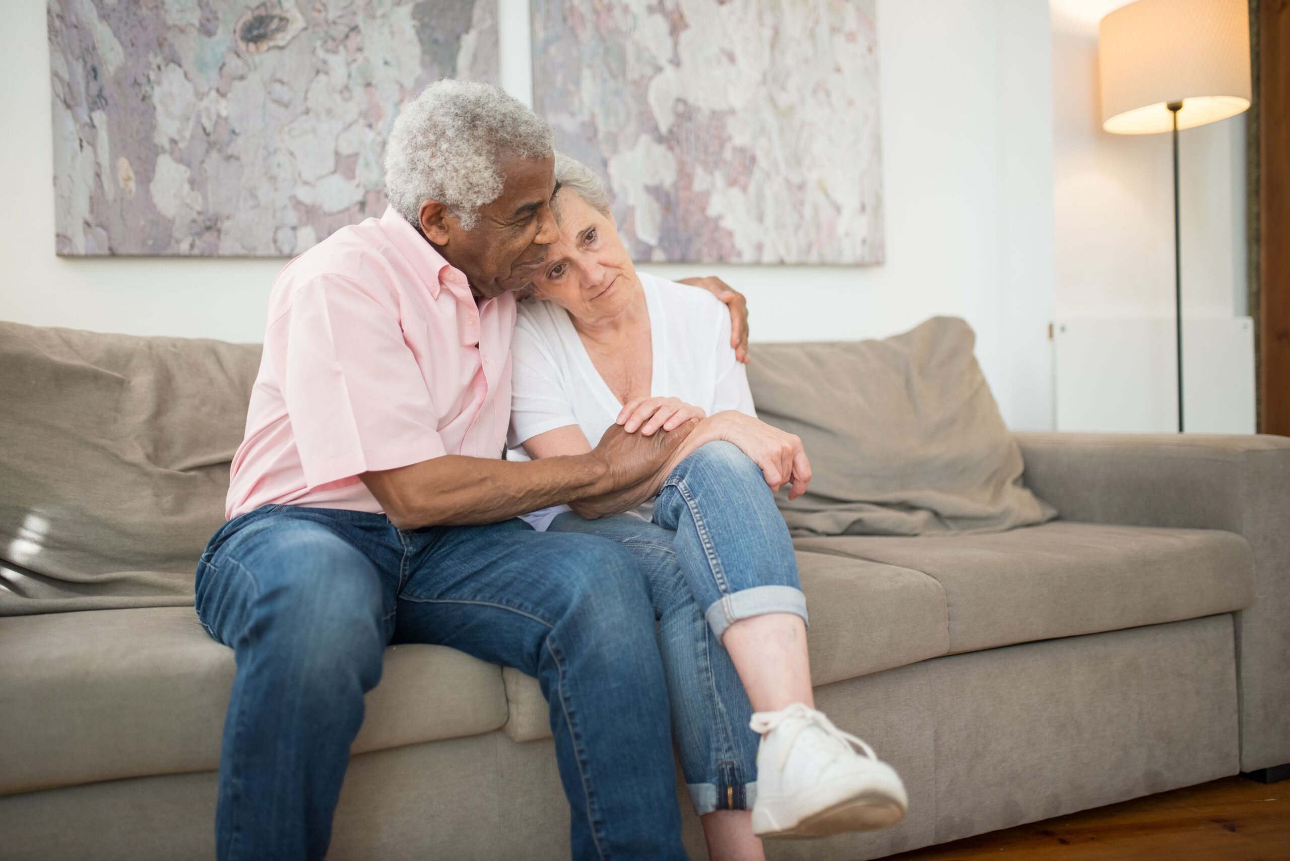 Elderly couple sitting close together on a sofa, sharing a tender moment with the man&rsquo;s arm around the woman.